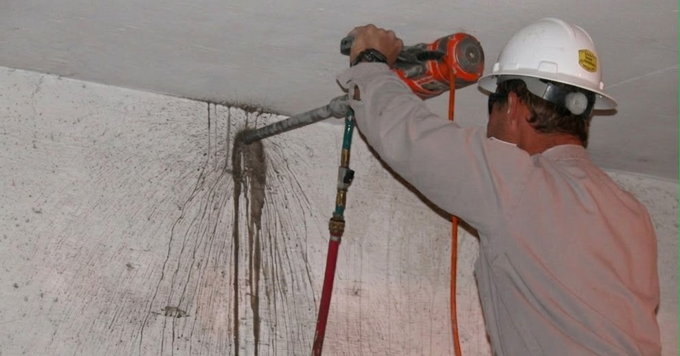 A construction worker wearing a hard hat drills into a concrete ceiling near a wall junction, demonstrating the high risk of hitting tendons in a post tension slab.